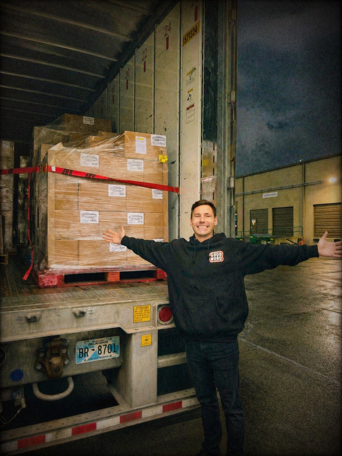Brad arms-out in front of a shrink-wrapped pallet of GOOD WORD cases loaded onto an Oklahoma-plated trailer