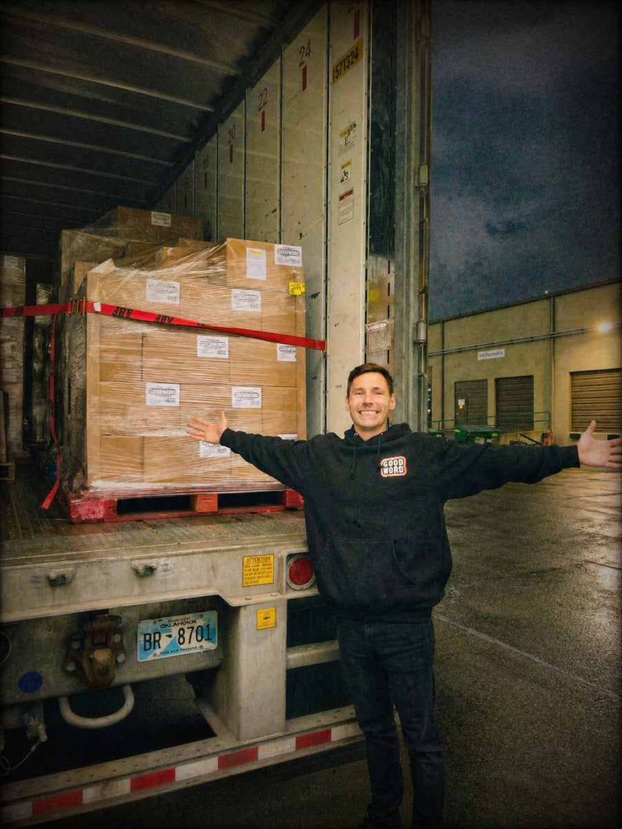 Brad arms-out in front of a shrink-wrapped pallet of GOOD WORD cases loaded onto an Oklahoma-plated trailer